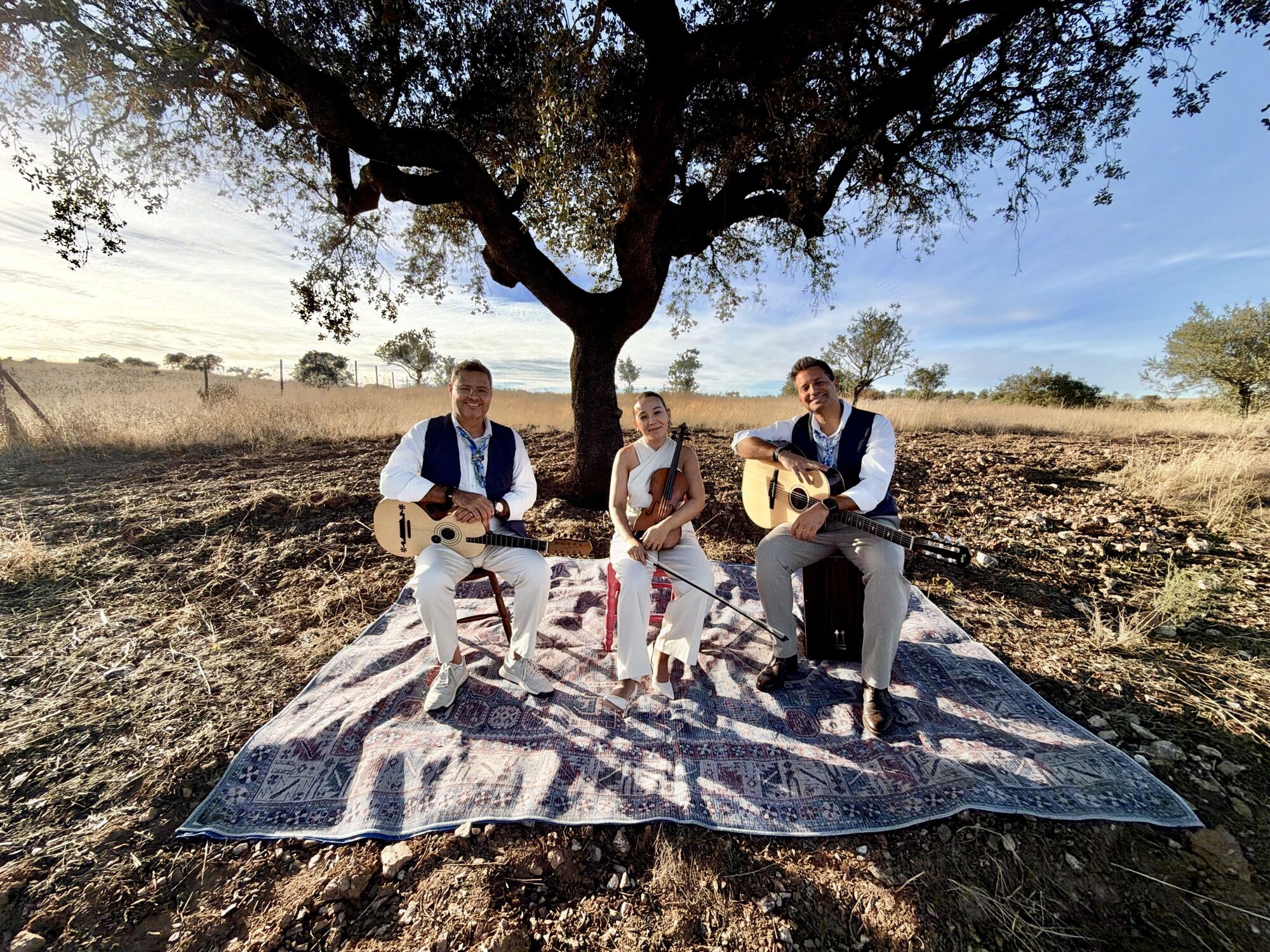 O trio Voz entre Cordas sentado sobre um tapete, à sombra de uma azinheira no Alentejo, com a viola campaniça, o violino e a guitarra